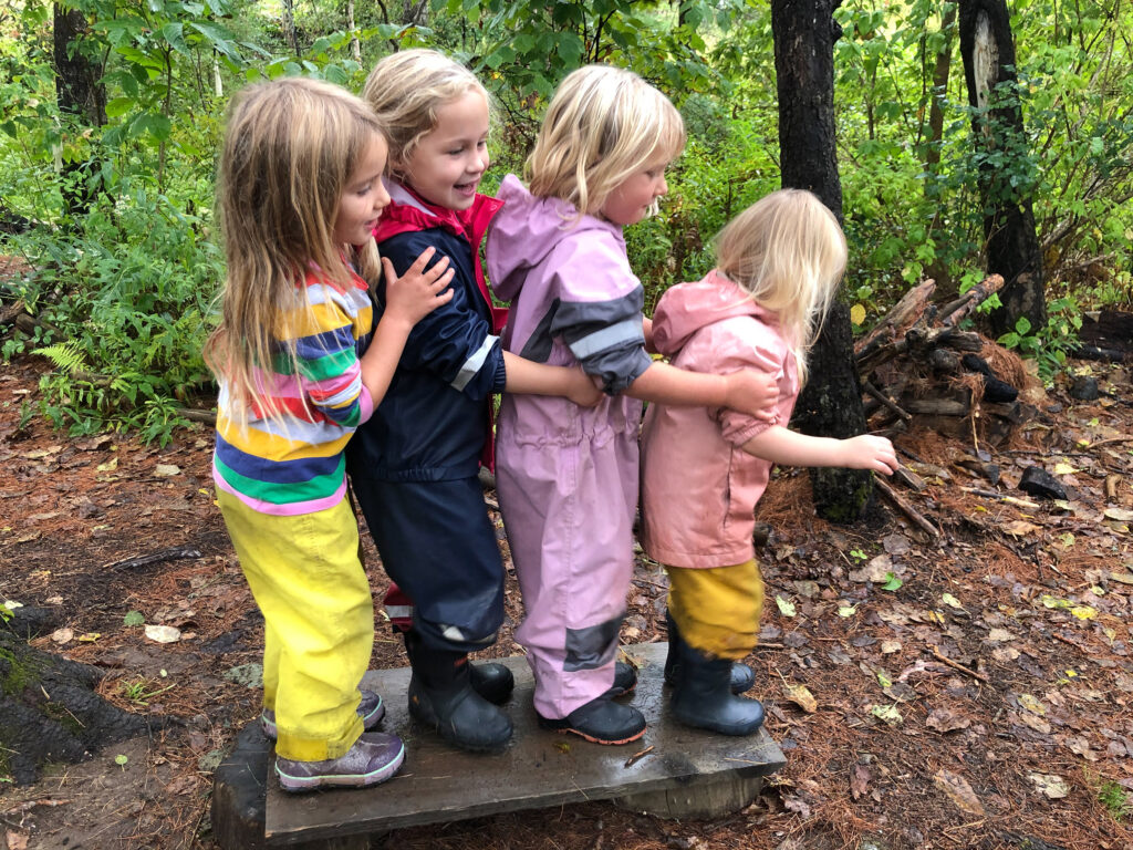 four preschoolers balance on a board in the woods in rainsuits